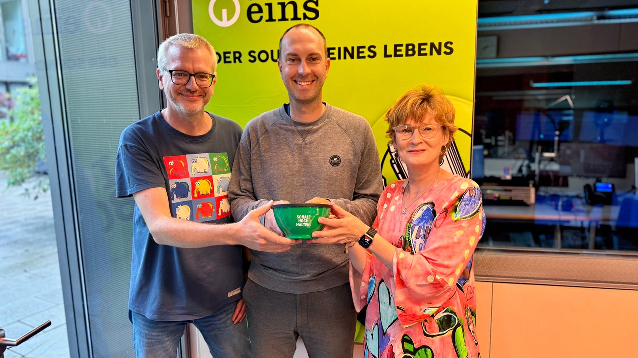 Anja Kwijas und JUK  mit Ole Werner im Bremen-Eins-Studio, die eine Müslischale hochhalten.