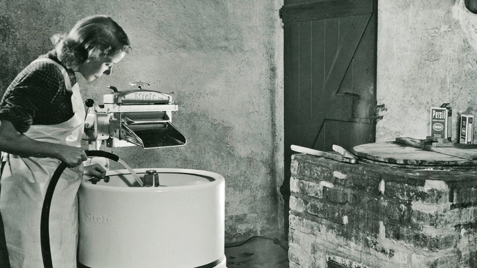 Eine Hausfrau mit Kittel steht im Waschkeller und befüllt eine Miele Waschmaschine in den 50er Jahren.