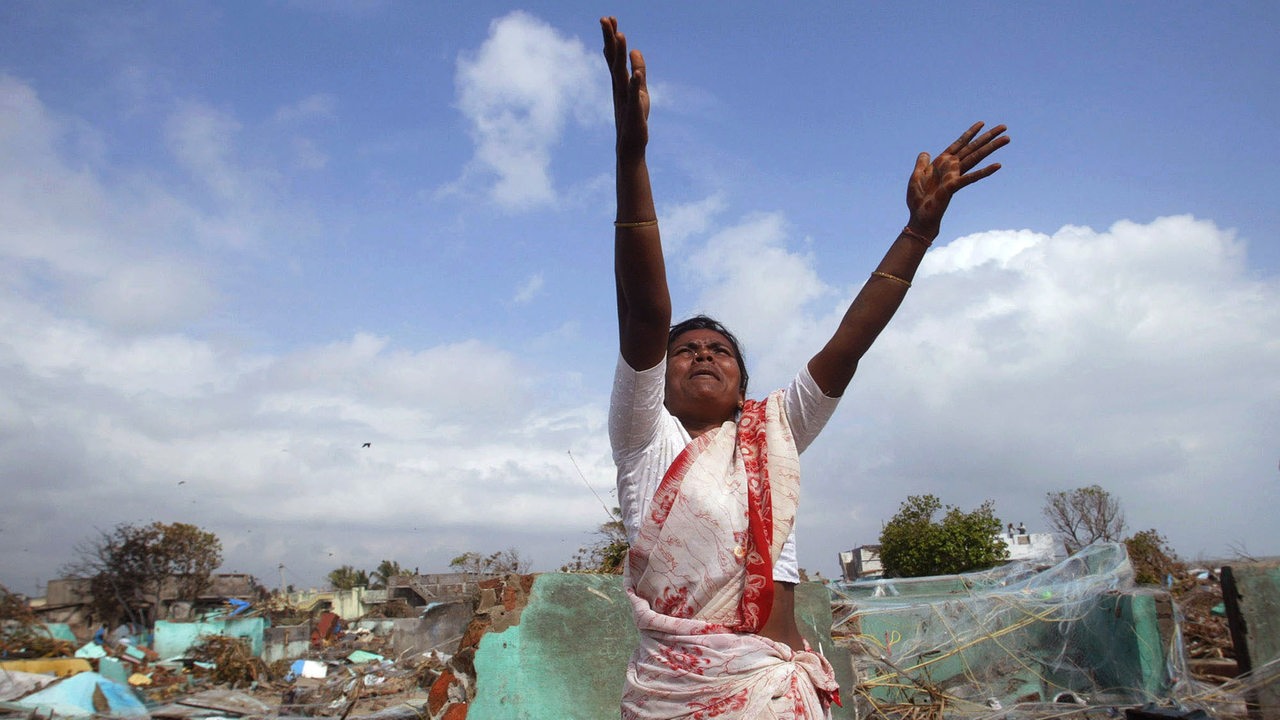 Eine indische Fischerfrau reckt hilfesuchend ihre Arme in die Höhe, um einen Hubschrauber zu erreichen. Hinter ihr die Trümmer nach dem Tsunami 2004