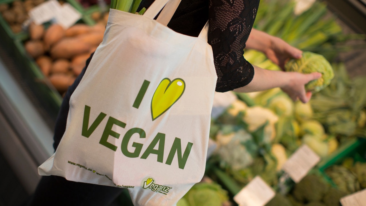 Eine junge Frau mit einem Leinenbeutel mit der Aufschrift "I love vegan" an einem Gemüsestand.